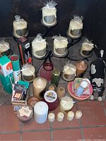 Multiple wrapped candles placed on black metal multi-arm candle holders, various other candles, matchboxes, and decorative items arranged on floor against dark background.