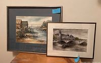 Two framed and matted art pieces displayed side by side on a table. One features a rustic dock scene, the other a cityscape with a bridge and boat.