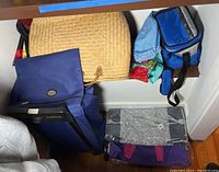 Multiple bags including the blue wheeled bag, straw bag, soft blue lunch bag, and other assorted bags stacked together.