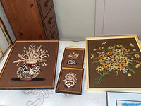 Four framed needlepoint stitched wall art pieces displayed on a surface, two large rectangular and two smaller rectangular frames with floral and vase designs.