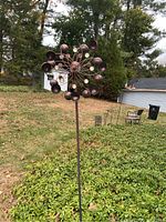 Full view of metal outdoor spinning art sculpture in a yard, showing the pole and circular design with petal-like elements.