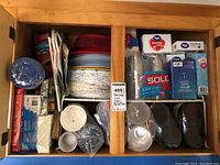 First photo showing various packed party supplies including paper plates, plastic cups, utensils, napkins, bags, and ground cover arranged in wooden cabinet compartments.