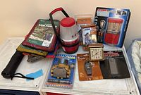 Photo showing emergency and camping supplies including a red Coleman lantern, folded rain ponchos, headlamp, clock, fire starter, whistle, and matches arranged on a surface.