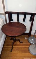 Photo of round wooden side table next to folding luggage rack in a room with hardwood floor.