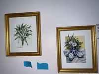Two signed floral paintings on mat, framed in gold, hung on a wall showing both lily of the valley and daisy flower themes