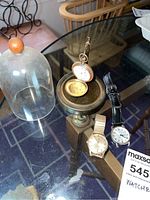 Three watches displayed on glass tabletop: pocket watch on brass stand with dome beside, Wittnauer wristwatch and silver-tone wristwatch