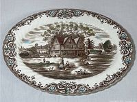 Full view of the oval platter showing the detailed colonial house and people scene with ornate floral borders in blue, brown, and green.