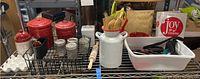 Wide view of shelf displaying red metal canisters, white ceramic items, wooden utensils, rolling pin, and plastic trays with various kitchen tools.