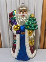 Full frontal image of a large mercury glass Santa Claus holiday figurine displaying colorful presents and a green Christmas tree with glitter accents.