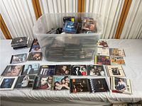 Wide view of a large plastic bin filled with dozens of CDs alongside several CDs spread out on a table showing album covers and cases.