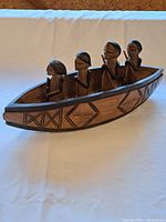 Full view of the wooden boat figurine with four men seated and standing inside, showing geometric carvings along the boat's sides.