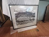 Framed print leaning against a wall with white backdrop and measuring tape showing size.