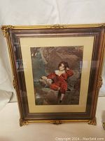 Front view of gilt-framed print showing full image of boy in red velvet outfit against rocky background, under glass, with cream matting and ornate gilt frame.