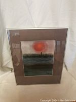 Framed watercolour painting showing sunset with red sun and dark landscape, surrounded by brown mat and white frame.