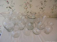 Photo showing 12 clear glass vases arranged on white surface against floral wallpaper background, highlighting variety in size and shape