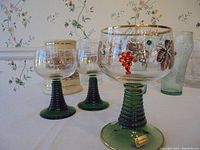 Photo showing decorative goblet-style glasses with green ribbed stems and gold grapevine motif on the bowl, with gold trim on the rim and base.