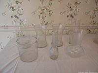 Eight clear glass vases displayed on white tablecloth against floral wallpaper, varying in size and shape including cylindrical and bulbous forms.