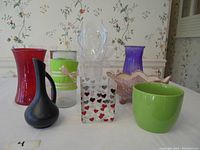 Nine assorted decorative vases and planters in different colors and shapes on a white tablecloth with floral wallpaper background