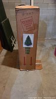 Image of original cardboard packaging box from Puelo's Manufacturing for the artificial tree labeled as fragile with a small photo of the tree on the box; box indicates 'Open This End'.