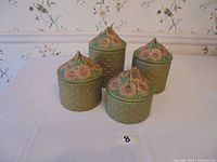 Four ceramic canisters with green basket-weave texture and pink floral lids on white cloth with floral wallpaper background.