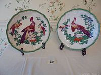 Both peacock-themed Chelsea House decorative charger plates displayed with stands, showing colorful bird illustrations and scalloped edges with green borders.