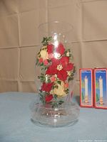 Hand painted hurricane candle holder with red and cream poinsettias floral design, clear glass with base, stood upright on a blue cloth background. Two boxed vintage electric window candles placed to the right of it.