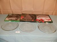 Three boxed and unboxed Christmas-themed glass platters displayed on a table, showing all items included in the lot