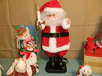 Front view of vintage lighted Santa Claus figure with candle, elf and snowmen trio, and several plush rabbits on cloth-covered table against beige backdrop.