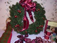 Photo of wreath hanging on wall, showing large burgundy velvet bow, red ball ornaments, pine cones, and fabric Advent doll leaning beside it. Ribbon rolls and Martha Stewart book partially visible.