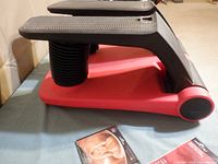 Side view of AirClimber stepper showing red base, black foot pedals, and instruction DVD on table.