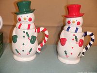 Two ceramic snowmen mugs with removable lids, one with green top hat and red striped handle, the other with red top hat and blue striped handle