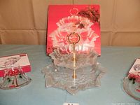Three Mikasa Christmas serving dishes displayed on a blue cloth backdrop, central two-tiered clear glass tidbit server with gold handle and scalloped edges, flanked by two smaller colored dishes