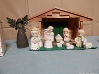 Close-up of wood framed nativity manger with 11 ceramic figures arranged in front, including Mary, Joseph, Baby Jesus, 3 Wise Men, Shepherd, two sheep, and a cow. A black angel statue stands behind the manger.