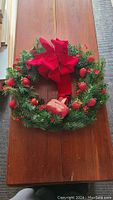 Decorative Christmas wreath laid flat on the floor, featuring a large red bow, red berries and balls, and a small basket attached.
