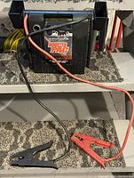 Photo of Booster Pac 25000 battery booster pack with red and black jumper cables laid out in front on a carpeted step surface.