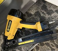 Yellow and black Craftex brad nailer placed on dark gray carpet, showing details of the nail magazine and air hose connector.