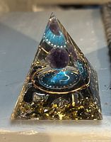 Photo showing small clear resin pyramid with blue circular element, purple stone, and gold and silver metallic flakes embedded
