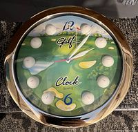 Photo of a round wall clock with a golf course scene on the face, golf balls marking the hours, gold bezel, and quartz movement indication.
