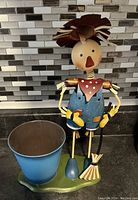 Photo features a blue metal planter pot next to a metal farmer figurine in blue overalls holding a broom, both set against a kitchen backsplash.