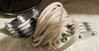 Photo of two metal triple-groove pulleys with shiny polished finish placed on a beige carpet, coiled white synthetic rope also shown intertwined with the pulleys.