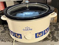 Full view of the Rival branded crock pot showing exterior design, control knob with settings Off, Low, High, Warm, and glass lid with silver handle.