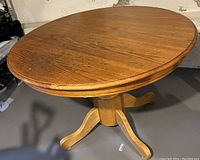 Round solid wood dining table with visible wood grain, single pedestal base with four legs, warm brown finish.