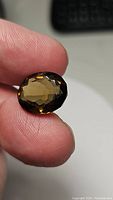 Close-up front view of the deep cognac colored oval cut Madeira citrine gemstone held between fingers showing facets and clarity.