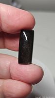 Photo of the golden sheen obsidian cabochon held between fingers showing its size, shape, and dark polished surface.