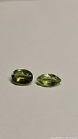 Two apple green peridot gemstones placed on a neutral background. One stone is oval cut and the other is marquise cut, showcasing their color and clarity.