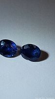 Close-up image of two oval cut blue sapphire gemstones showing their faceted surfaces and deep cornflower blue color under direct light.