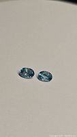 Two oval cut lagoon blue Cambodian zircon gemstones placed on a plain background, showing size and color attributes