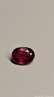 Oval cut blood red ruby gemstone placed on a light surface with visible facets and deep color.