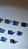 Eight square cut natural sapphires with blue and green tones arranged on a neutral background, showing color and cut details.