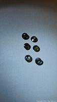 Six small round cut sapphires with green and blue color undertones visible in dim lighting on a plain background.